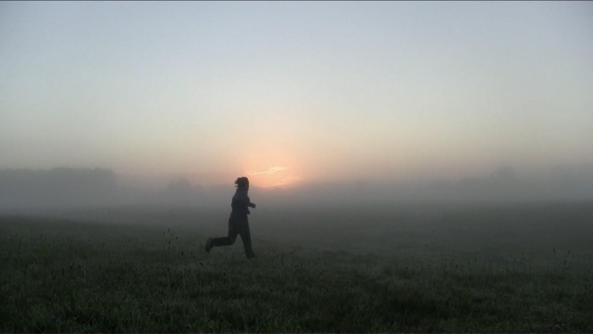 A person is jogging across a grassy field early in the morning. The landscape is covered in thick fog, blurring the horizon line.  We can see the red glow from a sunrise appearing amongst the misty background. The person running appears slightly mid air, in between steps. The sunrise makes them out like a silhouette on the land. The grass is covered in dew or frost.